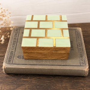 May include: A rectangular wooden box with a light green brick pattern and gold accents on the lid. The box sits atop an old book with the text "Modern Course in English". The box has a wooden base.