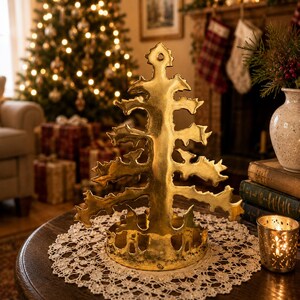 Può includere: Una decorazione per albero di Natale in metallo color oro è appoggiata su un tavolo rotondo di legno con un centrino di pizzo bianco. Una candela accesa in un portacandele dorato è accanto all'albero. Un albero di Natale con luci è sullo sfondo.