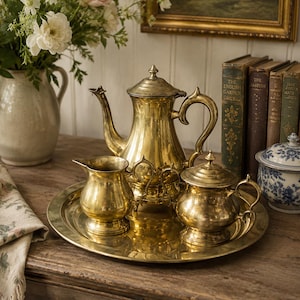 May include: A vintage brass tea set, including a teapot, creamer, sugar bowl, and serving tray. The set is displayed on a wooden surface, with a vase of flowers and antique books in the background. The tea set has a shiny, reflective surface.