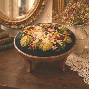 May include: A round footstool with a floral needlepoint design. The stool has a dark background with a colorful flower pattern and a wooden frame. The legs are curved. The stool is placed on a wooden surface.