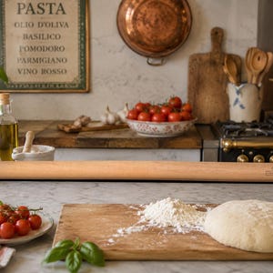 May include: A long, light-colored wooden rolling pin rests on a marble countertop, near a wooden cutting board with flour and dough. A plate of red cherry tomatoes and a basil leaf are in the foreground. The background includes kitchen items.