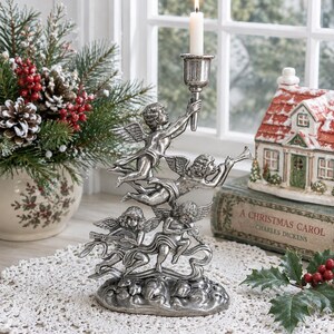 May include: A silver-colored angel candle holder with four cherubic figures holding a candle. The candle is lit, and the holder is on a white lace doily. A Christmas-themed arrangement is in the background.