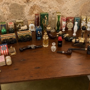 Pode incluir: Uma coleção de artigos vintage, incluindo frascos de perfume, caixas decorativas, cachimbos e estatuetas, dispostos numa mesa de madeira escura. Os artigos são diversos em forma, tamanho e cor, com materiais como vidro, metal e cerâmica.