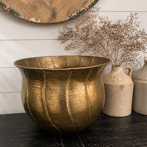 Op de afbeelding: Een grote, gehamerde messing plantenbak met een gegroefd ontwerp. Twee keramische kruiken en een vaas met gedroogde bloemen staan achter de plantenbak. De objecten staan op een zwart oppervlak, met een houten muur op de achtergrond.