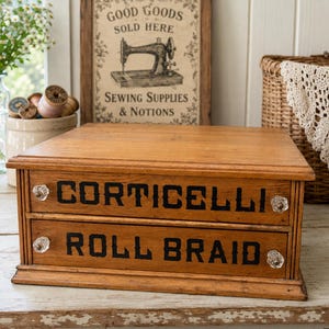 May include: Wooden sewing box with two drawers. The top drawer reads "CORTICELLI" and the bottom drawer reads "ROLL BRAID" in black lettering. The drawers have clear, crystal-like knobs. A vintage sewing supplies sign is in the background.
