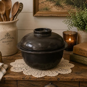 May include: A dark grey cooking pot with a lid rests on a cream lace doily. The pot is on a wooden surface, with a vintage container of wooden spoons and a small brass mister in the background. A lit candle and cookbooks are also visible.