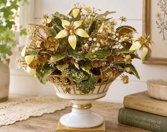 May include: A decorative floral arrangement in a white and gold ceramic vase. The arrangement features yellow and gold flowers with green leaves. The vase has a pedestal base and sits on a wooden surface, creating a vintage aesthetic.