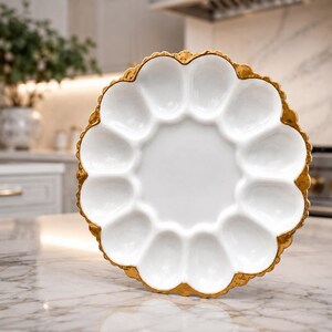 Peut inclure: Plat à servir en céramique blanche avec bord festonné et bordure dorée. Le plat comporte un cercle central et 12 compartiments individuels, idéal pour servir des amuse-gueules ou des collations.