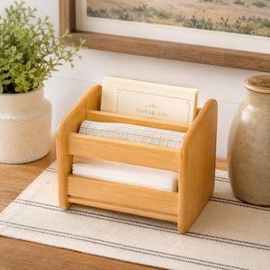 May include: A light-colored wooden organizer with three compartments. The top section holds a card that reads "THANK YOU." The middle section contains a folded cloth, and the bottom section holds white paper. The organizer sits on a striped surface.