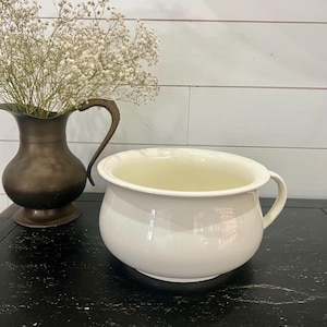 May include: A white, antique chamber pot with a handle. Beside it, a bronze-colored pitcher holds a bouquet of white flowers. The objects sit on a black surface against a white-paneled wall. The chamber pot is a classic piece.