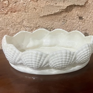 Könnte beinhalten: Eine weiße, muschelförmige Keramikschale mit gewelltem Rand. Die Schale hat eine Reihe von geprägten Muscheln um den Sockel. Die Schale steht auf einer dunkelbraunen Oberfläche.