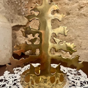 Könnte beinhalten: Ein messingfarbener Weihnachtsbaum-Kerzenhalter. Der Baum hat ein detailliertes Ausschnittdesign mit Ästen und einem stabilen Sockel. Der Kerzenhalter steht auf einer weißen Spitzendecke.