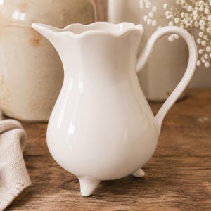 May include: A white ceramic pitcher with a bulbous body and three small feet. The pitcher has a curved handle and a scalloped rim. The background includes other ceramic items and a wooden surface.