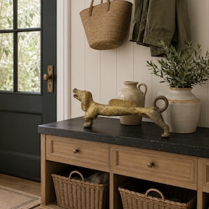 May include: A gold-colored dachshund statue is displayed on a wooden console table with two wicker baskets. A woven tote bag hangs on the wall above, along with a green jacket. A vase with greenery and a ceramic vase are also on the table.