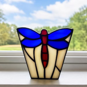 May include: Stained glass dragonfly-shaped candle holder. The dragonfly has blue wings, a red body, and is set against a cream-colored background. The candle holder is outlined in black and sits on a white windowsill.