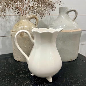 Op de afbeelding: Een witte keramische kan met een geschulpte rand en drie kleine voetjes. Achter de kan staan twee antieke steengoed kruiken en een vaas met gedroogde bloemstelen. De objecten staan op een zwart oppervlak tegen een wit geschilderde houten achtergrond.