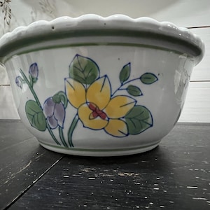 May include: A white ceramic planter with a scalloped rim and a floral design featuring yellow, green, and purple flowers.