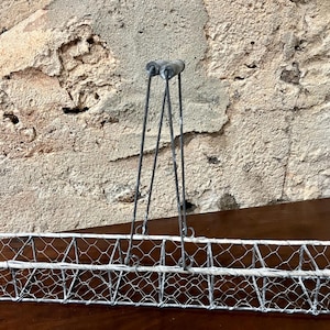 Könnte beinhalten: Ein rechteckiger Drahtkorb mit Griff. Der Korb besteht aus silberfarbenem Drahtgeflecht und hat eine lange, schmale Form. Der Griff besteht aus demselben Draht und ist mit zwei vertikalen Stützen am Korb befestigt. Der Korb wird wahrscheinlich zur Aufbewahrung oder Präsentation verwendet.