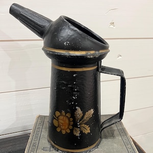 Könnte beinhalten: Schwarze Öldose aus Metall mit einem goldenen, bemalten Blumendesign und einem Ausguss. Die Dose hat einen Griff und steht auf einem Stapel Bücher.