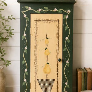 May include: A dark green wooden cabinet with a light-colored door featuring a painted pear design. The cabinet is adorned with floral vine accents and a black knob. A vase of flowers sits on top.