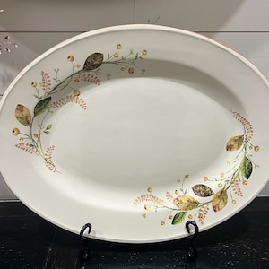 Puede incluir: Plato de servir ovalado blanco con un borde decorativo de hojas verdes, hojas marrones y pequeñas flores rosas y amarillas.