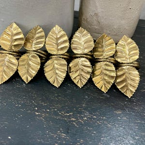 May include: A set of ten gold-colored leaf-shaped napkin rings. Each ring features a detailed leaf design with visible veins. The rings are arranged in two rows of five, showcasing a decorative table setting element.