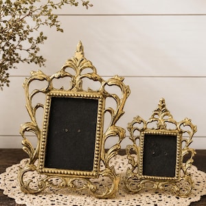 May include: Two gold-colored metal picture frames with ornate, scrolling filigree borders and beaded inner edges. Each frame features a rectangular black insert and a pointed top finial, showcasing a detailed, metallic finish with intricate curves.