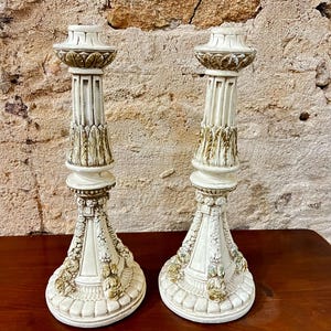 Op de afbeelding: Twee crèmekleurige decoratieve kandelaars met sierlijke details. De kandelaars hebben een kolomontwerp met gouden en groene accenten. De bases zijn rond en versierd met bloemen- en bladerpatronen. De kandelaars zijn ongeveer 30 cm hoog.