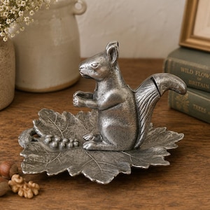 May include: A silver-colored squirrel nutcracker sits atop a leaf-shaped dish. The squirrel is detailed with textured fur and holds its paws up. The dish has a detailed leaf design. Walnuts and hazelnuts are scattered nearby. Decorative home decor.