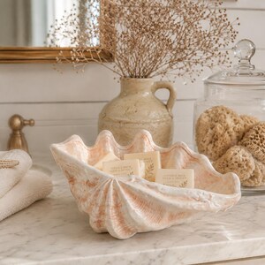 Pode incluir: Uma saboneteira decorativa em forma de concha num ambiente de casa de banho. A saboneteira é branca com detalhes em rosa e contém vários sabonetes pequenos. Um frasco de vidro com esponjas, um vaso com flores secas e uma torneira dourada também são visíveis.