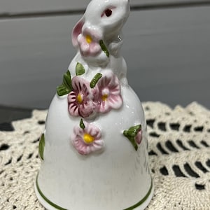 May include: White ceramic bell with a rabbit figurine on top. The rabbit is surrounded by pink and green flowers.