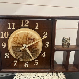 May include: A brown wooden wall clock with a white face and black numbers. The clock has a small shelf above it with a gold owl figurine. The clock face has a picture of a dried flower arrangement.
