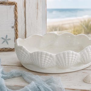 Puede incluir: Un cuenco de cerámica blanca con forma de concha, con un borde festoneado. El cuenco está sobre una superficie de madera blanca, con un letrero que dice "by the sea" en el fondo. También se ven una estrella de mar y conchas.