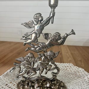 May include: Silver-toned candle holder featuring three cherubs playing musical instruments and one holding a torch. The cherubs have detailed wings and are set on a decorative base. The candle holder is a decorative home accent.