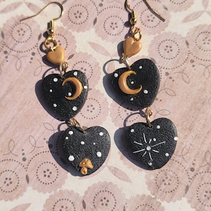 Puede incluir: Un par de pendientes colgantes con tres abalorios en forma de corazón negro. Cada abalorio tiene un diseño diferente: una luna creciente, un hongo y una estrella. Los abalorios están conectados por anillos de salto dorados. Los pendientes están hechos de arcilla polimérica y tienen ganchos dorados.