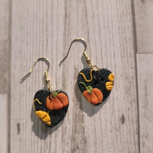 Puede incluir: Un par de pendientes en forma de corazón con fondo negro y calabazas naranjas y hojas amarillas. Los pendientes están hechos de arcilla polimérica y tienen ganchos dorados.