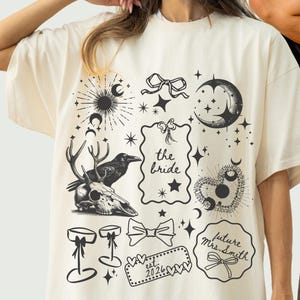May include: Two oversized t-shirts, one ivory and one grey, with a black and white graphic print. The design includes a deer skull, raven, moon, stars, and text. The ivory shirt says "the bride". The grey shirt says "bride's court".