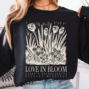 May include: Black sweatshirt with an ivory floral design and the text "LOVE IN BLOOM." The text "Gabby's Bachelorette Holland, Michigan" is also included. The sweatshirt has long sleeves.