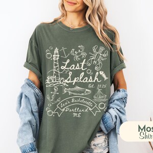 May include: A moss green t-shirt with a white graphic design. The design includes a lighthouse, fish, lobster, crab, and the words "Last Splash" and "Lexi's Bachelorette Portland ME". The shirt is a relaxed fit.