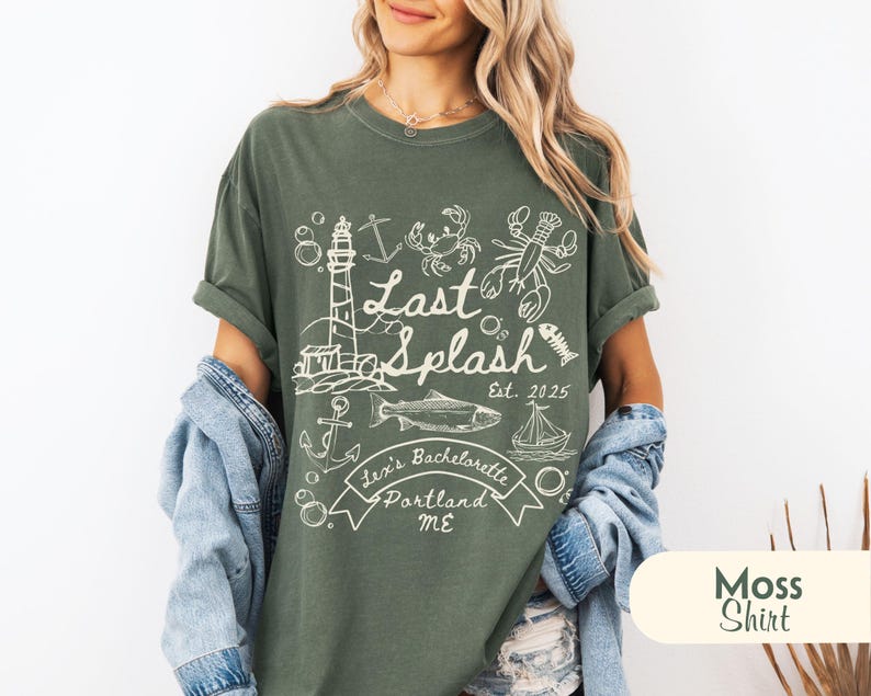 May include: A moss green t-shirt with a white graphic design. The design includes a lighthouse, fish, lobster, crab, and the words "Last Splash" and "Lexi's Bachelorette Portland ME". The shirt is a relaxed fit.