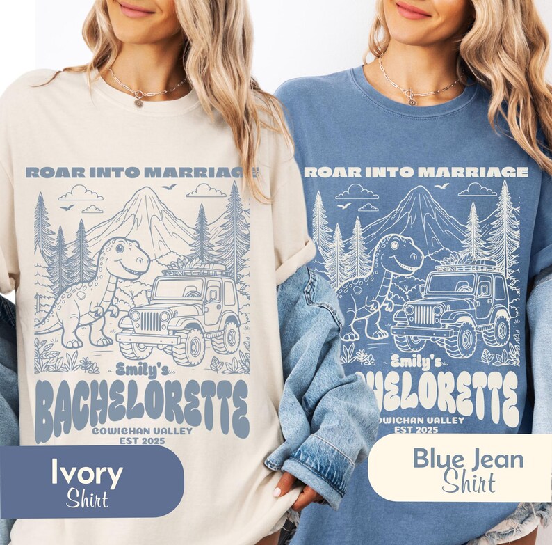 Op de afbeelding: Twee T-shirts met ronde hals, &eacute;&eacute;n ivoorkleurig en &eacute;&eacute;n blauw, met een grafische print van een dinosaurus en een terreinwagen in een bergbos. De tekst luidt ROAR INTO MARRIAGE, Emily's BACHELORETTE, COWICHAN VALLEY en EST 2025 in blauwe inkt.