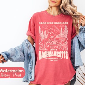 Op de afbeelding: Een watermeloenkleurig t-shirt met een ivoorkleurige grafische print van een dinosaurus, een jeep en bergen. De tekst luidt ROAR INTO MARRIAGE, Carol's BACHELORETTE, COWICHAN VALLEY en EST 2026. Het t-shirt wordt gedragen met een lichtblauw spijkerjack.