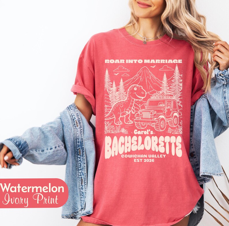 Op de afbeelding: Een watermeloenkleurig t-shirt met een ivoorkleurige grafische print van een dinosaurus, een jeep en bergen. De tekst luidt ROAR INTO MARRIAGE, Carol's BACHELORETTE, COWICHAN VALLEY en EST 2026. Het t-shirt wordt gedragen met een lichtblauw spijkerjack.