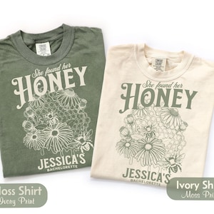 May include: A moss green t-shirt with ivory print and an ivory t-shirt with moss green print. Both feature a graphic of a honeycomb, flowers, and bees with the text "She found her HONEY" and "JESSICA'S BACHELORETTE" printed on the front.