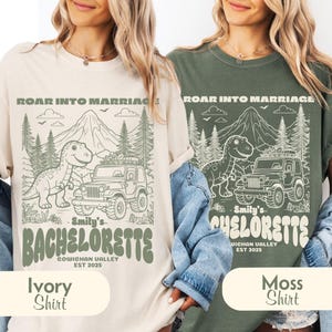 Op de afbeelding: Een ivoorkleurig en een mosgroen t-shirt, elk met een grafische print van een dinosaurus, een terreinwagen, bergen en bomen. De tekst luidt ROAR INTO MARRIAGE, Emily's BACHELORETTE, COWICHAN VALLEY en EST 2025 in een retro lettertype.