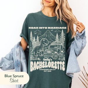 Op de afbeelding: Een donkergroen t-shirt met een witte grafische print van een dinosaurus, een jeep en bergen. De tekst luidt ROAR INTO MARRIAGE, Emily's BACHELORETTE, COWICHAN VALLEY, EST 2025. Het t-shirt wordt gedragen onder een lichtblauw spijkerjack.