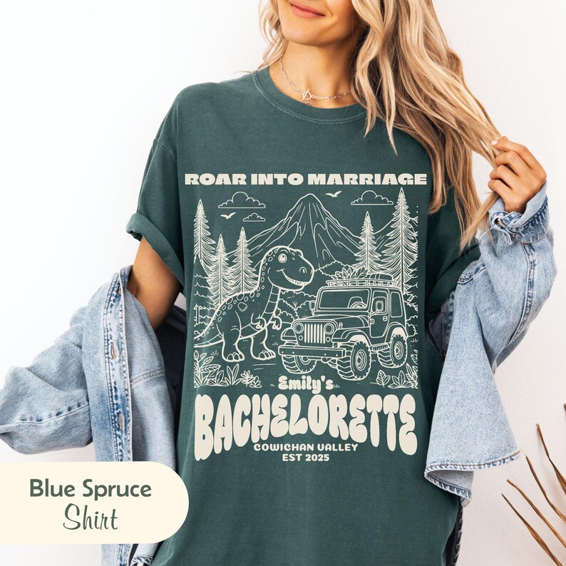 Op de afbeelding: Een donkergroen t-shirt met een witte grafische print van een dinosaurus, een jeep en bergen. De tekst luidt ROAR INTO MARRIAGE, Emily's BACHELORETTE, COWICHAN VALLEY, EST 2025. Het t-shirt wordt gedragen onder een lichtblauw spijkerjack.