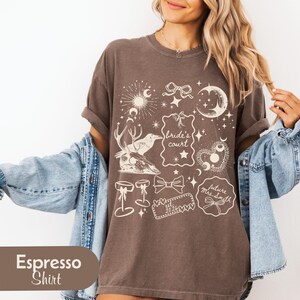 Pu&ograve; includere: T-shirt color espresso con illustrazioni fantasiose color crema. Il design include elementi celesti, fiocchi e il testo "Bride's Court" e "future Mrs. Smith est. 2024".