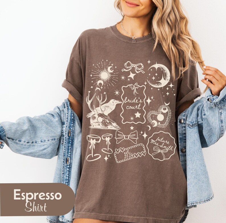 Pu&ograve; includere: T-shirt color espresso con illustrazioni fantasiose color crema. Il design include elementi celesti, fiocchi e il testo "Bride's Court" e "future Mrs. Smith est. 2024".