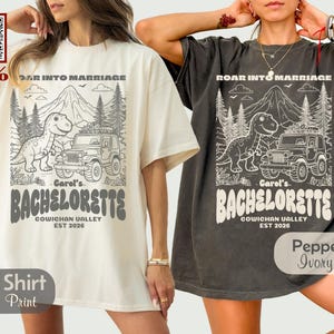 Op de afbeelding: Twee oversized t-shirts, &eacute;&eacute;n ivoorkleurig en &eacute;&eacute;n antracietgrijs, elk met een grafische print van een dinosaurus, een jeep, bergen en dennenbomen. De tekst luidt ROAR INTO MARRIAGE, Carol's BACHELORETTE, COWICHAN VALLEY en EST 2026.